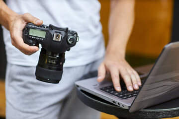 A young man stands outdoors with a camera and takes off a photo on a laptop.