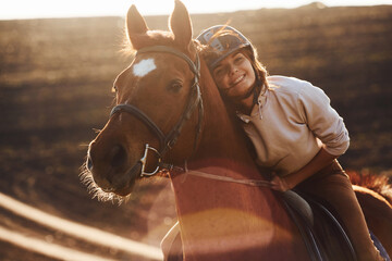 Young woman in protective hat with her horse in agriculture field at sunny daytime