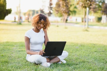 Young attractive dark-skinned college student wearing tank top and jeans sitting on the lawn at campus on sunny day, working on her thesis using laptop computer, looking busy and concentrated