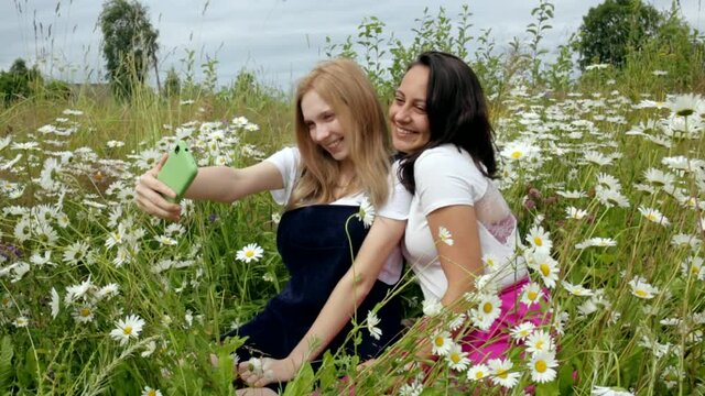 Two happy young blogger girls greet their subscribers on the phone in white t-shirts, take photos on the phone, laugh and inhale the scent of wild meadow flowers while enjoying a summer day. Chamomile
