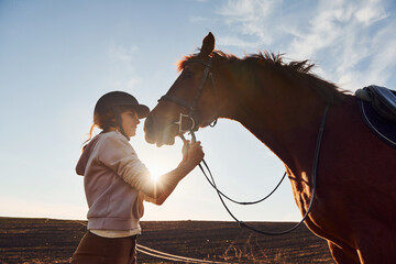 Young woman standing with her horse in agriculture field at sunny daytime