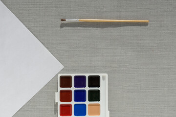 Brush, pallet watercolor paint and empty sheet of paper on grey background. no people. Top view. copy space. Workplace artist. drawing kit. fine art, creativity, painting and artistic tools concept