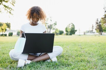Thoughtful cute mixed female international student with curly hair is sitting on fresh grass with modern laptop in public park, leaning on apple tree and wistfully looking aside during her break
