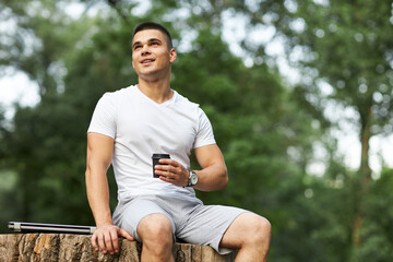 A young man sits in the park on a large stump with coffee in his hands.