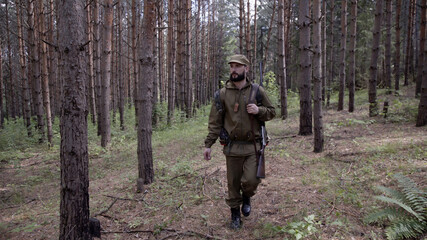 Hunting hunter men with gun walking through forest during hunting season. Man hunter outdoor in forest hunting alone. He has backpack and comfortable camouflage clothes.