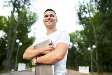Fototapeta premium A young man stands in the park with a laptop tightly clasped to his chest. He looks straight.