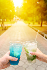Male and female hands hold plastic glasses with iced fruit lemonades in a city park. Refreshing non-alcoholic cocktails. Summer sunny day. Vertical orientation, copy space.