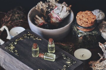 Small essential oil bottles placed on dark brown wooden witch's box. Closeup with various dried herbs, plants, flowers, sage smudge stick, candles in the blurred background. Messy wiccan witch altar
