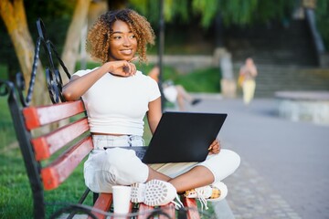 Cheerful young black girl on a wooden bench in a park is using the laptop; dazzling smiling Brazilian female outdoors on the bench with the netbook in hands and small leather bag on the left