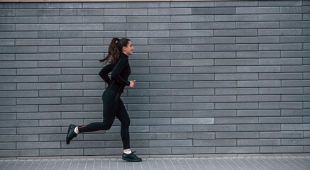 Young sportive girl in black sportswear running outdoors near gray wall
