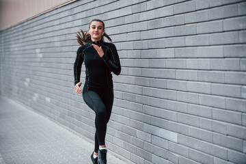 Young sportive girl in black sportswear running outdoors near gray wall