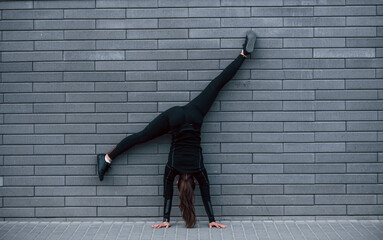 Young sportive girl in black sportswear doing hard handstand exercises outdoors near gray wall