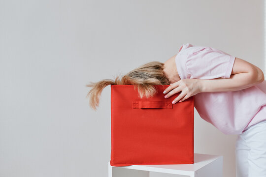 The Girl Hid Her Face And Half Her Head In A Bright Red Box. The Girl Bent Down To Hide Her Face And Head In A Box Against A Light Plain Gray Background