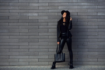 Fototapeta na wymiar Beautiful brunette with curly hair and in black clothes holding bag outdoors near wall
