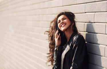Fototapeta premium Potrait of cheerful brunette with curly hair and in black clothes leaning on the wall and talks by phone