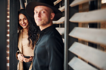 Woman with black curly hair and her man standing together near wooden windows