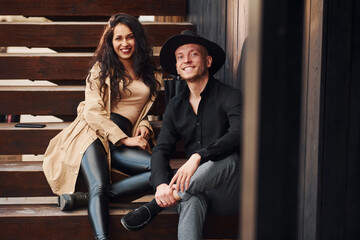 Fototapeta na wymiar Cheerful brunette in black leggings sitting on wooden stairs with her man in hat