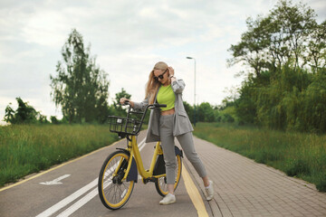 Young model girl in modish clothes posing with yellow bicycle at the bike path
