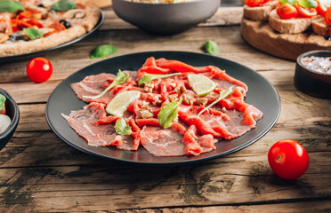 Black plate with carpaccio of beef with baby arugul and lime on rustic wooden background. Top view