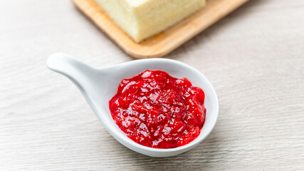 Strawberry Jam in white ceramic cup placed on wooden table with blurred bread background. Food close up