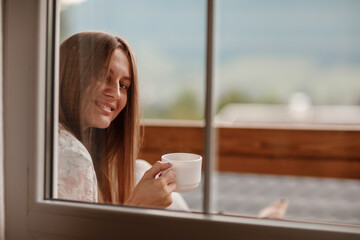 Young female standing after taking a shower in the morning on balcony of the hotel. holding a cup of coffee or tea in her hands. Looking outside nature forest and Mountain