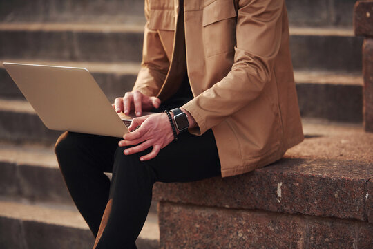 Sitting on stairs with laptop on legs. Elegant young man in formal classy clothes outdoors in the city