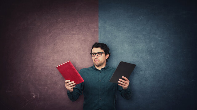 Pensive male student chooses between a traditional book and electronic PC tablet over a split wall with red and blue sides. Future education concept. Doubtful reader makes a difficult choice.