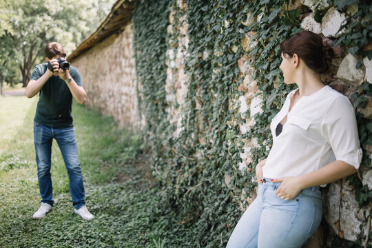 Photographer shooting woman leaning against wall with ivy. Man with professional camera taking pictures of woman who is posing on old wall.