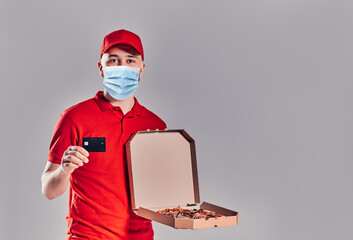Young bearded delivery man in red uniform in protective medical mask with pizza and credit card isolated on gray background.
