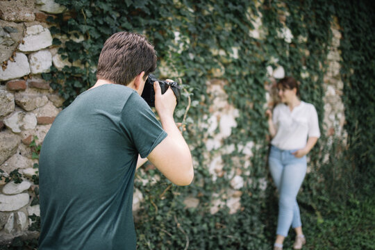 Back view of photographer taking photos of woman outdoor. Back of man using professional camera and taking pictures of blurred girl standing next to wall covered with ivy.