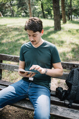 Obraz premium Portrait of young man using tablet outdoor. Photographer sitting next to camera on bench in park while using tablet.