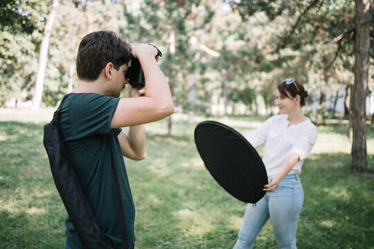 Man with camera taking photos of blurred girl with reflector. Side view of photographer taking pictures in park of out of focus woman who is holding collapsible reflector.