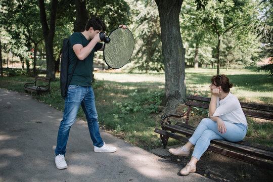 Photographer holding camera and reflector taking photos in park. Male photographer taking photos of girl who is sitting on wooden bench in park.