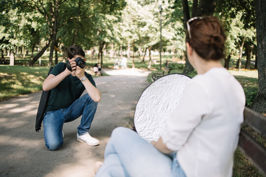 Front view of photographer taking pictures of blurred model. Squatting man with professional camera photo shooting female model with reflector in park.