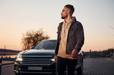 Handsome unshaved man in fashionable clothes walks near his black car at evening time