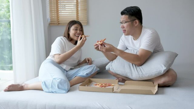 Normal shot : Young attractive happy asian married couple eat pizza, take-out food or fast food on bed feeling positive and relax on weekend quarantine lifestyle activity.