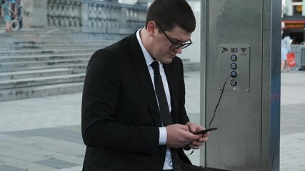 Confident businessman in black formal suit at bus stop puts phone on charge and talks on it. New...