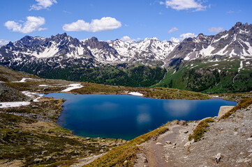 View of a deep blue high-altitude lake. In the foreground, a hiking trail and in the background a snow-covered mountain range.