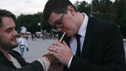 Hipster gives a light to a serious businessman in a black formal suit and glasses. Lung cancer concept