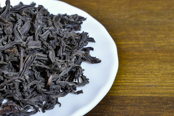 Large-leaf black tea on a white plate on a brown wooden background. Close-up Dried large black tea...