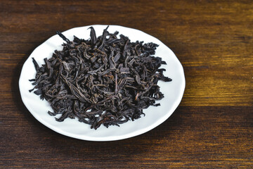 Large-leaf black tea on a white plate on a brown wooden background. Close-up Dried large black tea leaves in a white saucer on a wooden surface. Selective focus. 