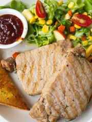 closeup of pork steak with salad, bread and barbecue sauce on a white plate