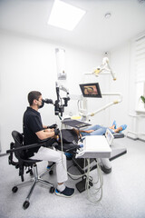 Delightful girl with patient bib on a dental chair and a dentist who sits next to her looks on her teeth using a dental microscope and holds a dental bur and a mirror.