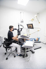 Delightful girl with patient bib on a dental chair and a dentist who sits next to her looks on her teeth using a dental microscope and holds a dental bur and a mirror.