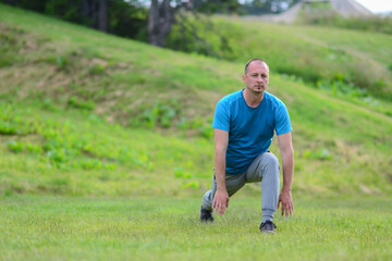 Fit man doing stretching exercises outdoors, middle age male sportsman stretching and preparing to run, attractive adult runner stretching the legs standing on grass in the nature, sports fitness