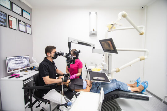 Male dentist and two female assistants treating patient teeth with dental tools - microscope, mirror and drill at dental clinic office. Medicine, dentistry and health care concept. Dental equipment