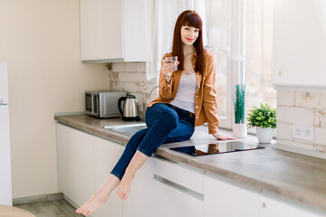 Attractive young woman in denim jeans and mustard casual shirt, sitting with glass of drinking water on the kitchen countertop in modern light home kitchen. Healthy lifestyle concept