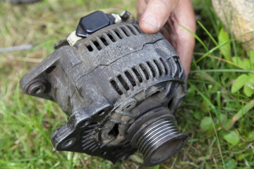 Used car alternator with a V-shaped pulley close up in hand on green grass background, outdoor vehicle electric equipment repair