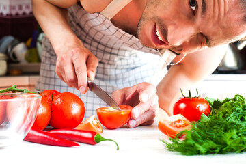 Young man with a knife cutting a fresh ripe tomato, smiling and winking. Husband is preparing a lunch. Healthy food concept. Close-up.