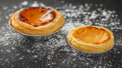 two sizes of sweet egg tarts vanilla custard and caramel with flour on dark grey texture background with copy space, 64 x 27 ultra widescreen aspect ratio